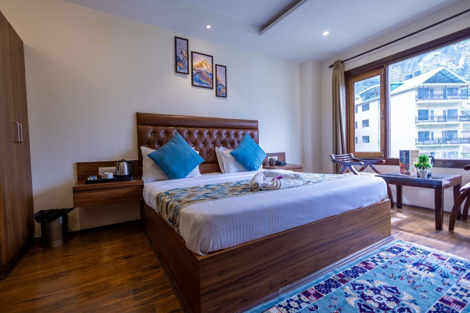 A hotel room with a large bed, wooden headboard, two nightstands, a desk, and a view of a building through a window.