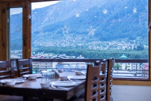 Uma sala de jantar com mesa posta para dois, com vista para uma cidade montanhosa, com edifícios e vegetação.