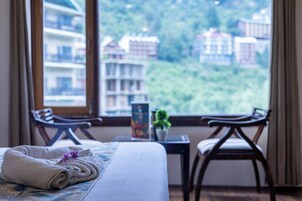 Cama bem arrumada com uma toalha enrolada e uma flor roxa, duas cadeiras de madeira, uma mesa com uma planta e vista para edifícios pela janela.