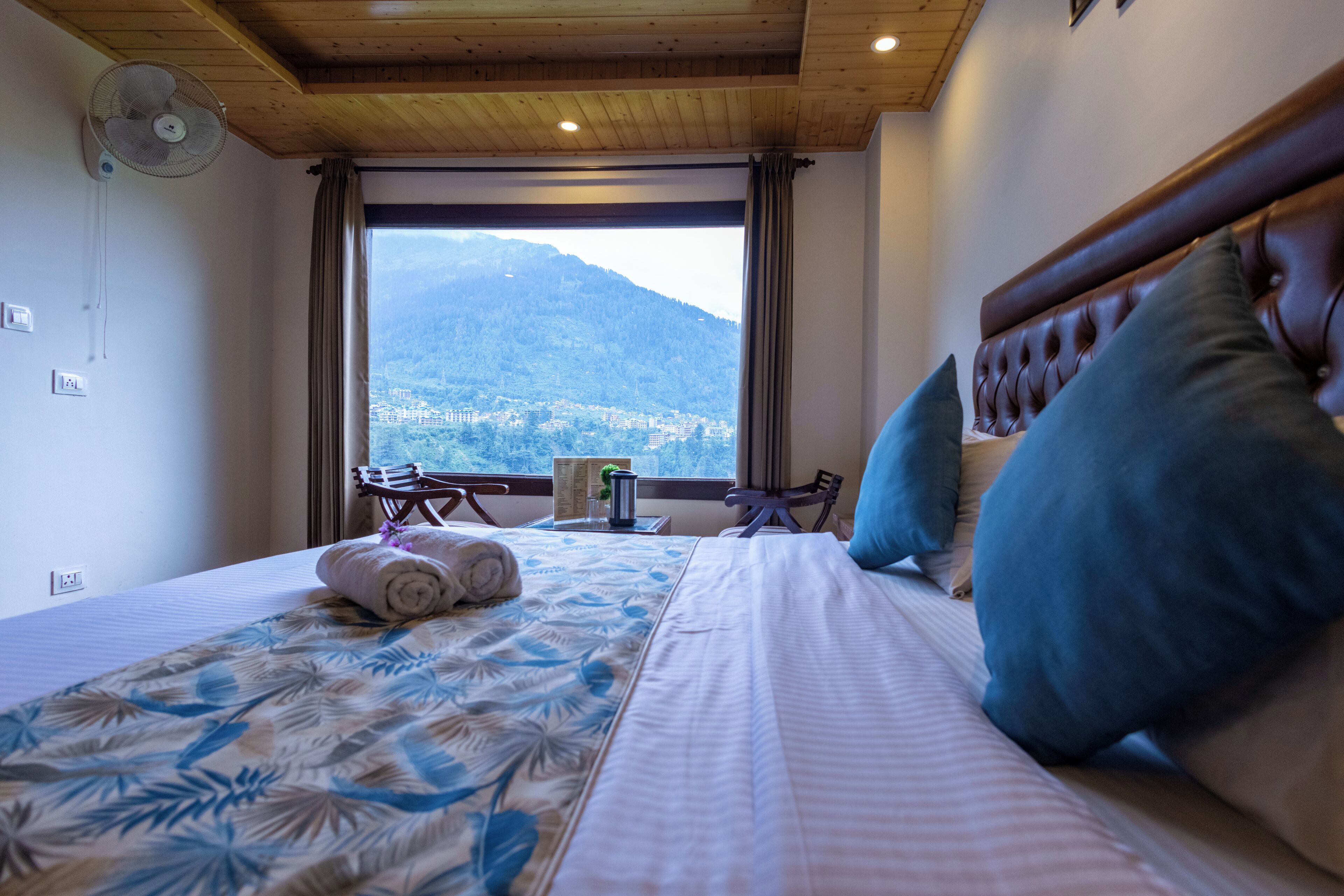 A hotel room with a bed, a fan, a window with a mountain view, and a table with a chair.