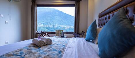 A hotel room with a bed, a fan, a window with a mountain view, and a table with a chair.