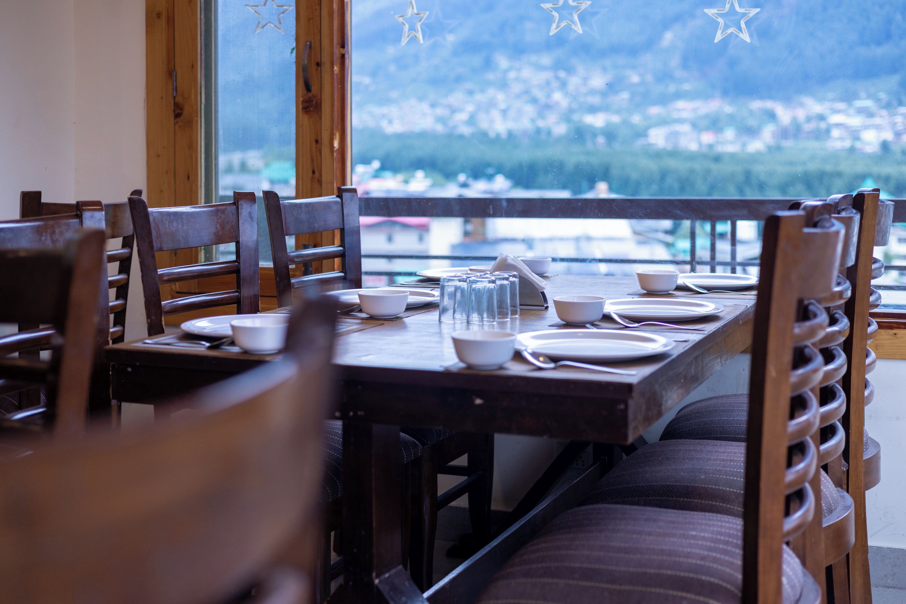 Mesa lista con platos, tazones y vasos, con vistas a un paisaje de un pueblo y montañas.