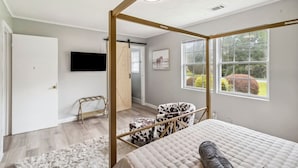 Un dormitorio con cama con dosel, televisor montado, ventana con vistas a zonas verdes y puerta que da a otra habitación.