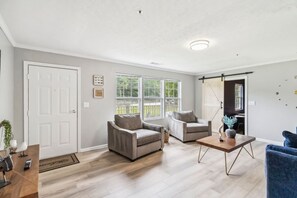 A spacious living room with a large window, a door, and a blue chair. - 5BR with Game Room & Huge Backyard (Lawrenceville)