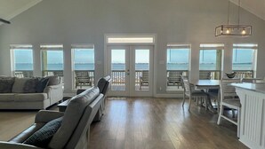 A spacious living room with a large window overlooking the sea, a dining area with white furniture, and a kitchen with a white countertop. - Bay Views at The Anchor Holds - formally Overboard (Grand Isle)