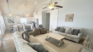 A spacious living room with a large sectional sofa, a smaller sofa, and a coffee table. There is a dining area with a table and chairs, and a kitchen in the background. - Bay Views at The Anchor Holds - formally Overboard (Grand Isle)