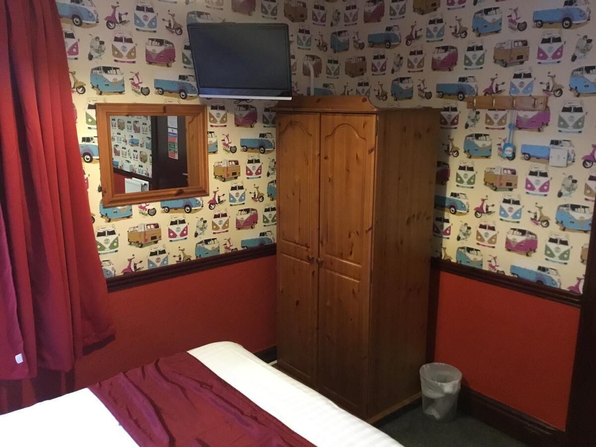 A bedroom with a bed, a wooden wardrobe, a mirror, a television on the wall, and a red curtain.