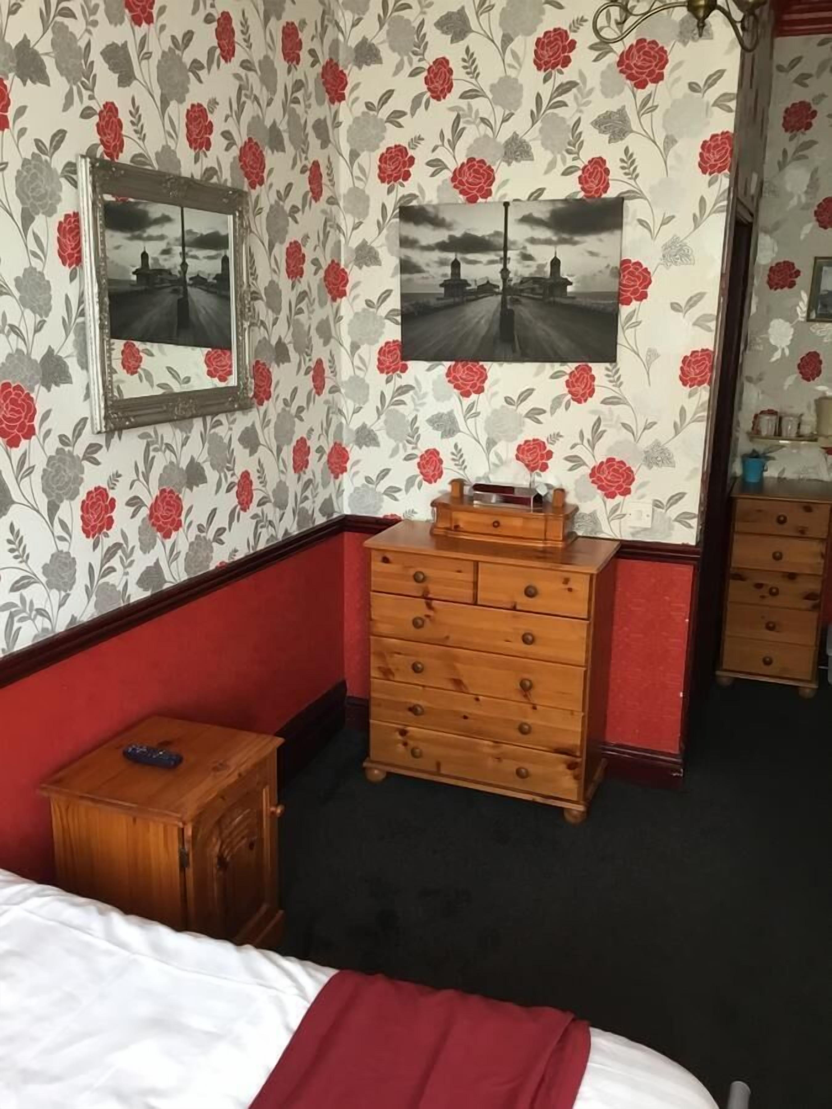Ein Schlafzimmer mit floraler Tapete, einem hölzernen Schminktisch, einem Spiegel und einem Bett mit roter Decke.