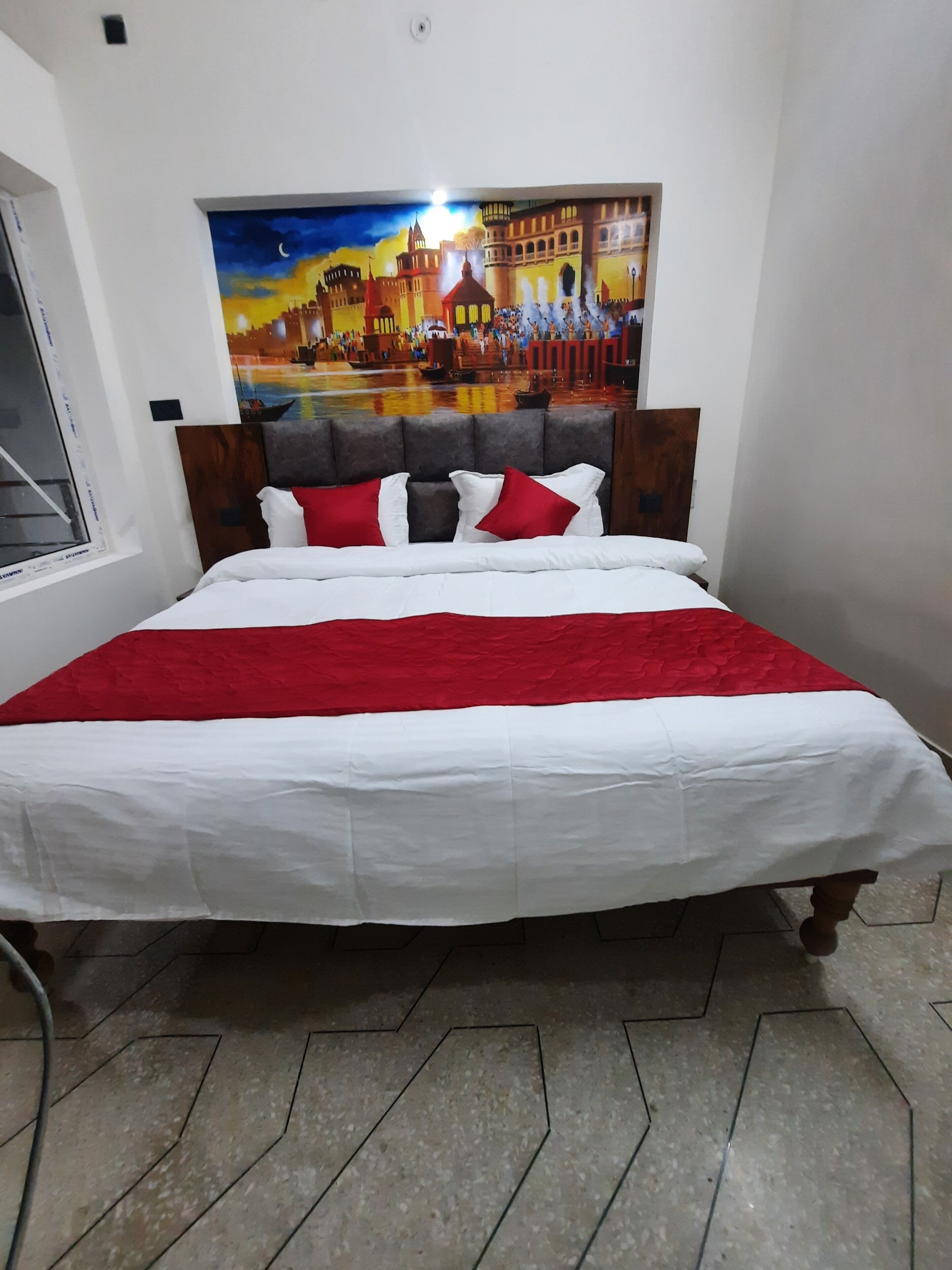 Una cama con una colcha blanca y roja, dos mesitas de noche de madera y un cuadro de un paisaje urbano en la pared.