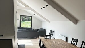 Un espace repas moderne avec une longue table en bois et des chaises noires, un banc intégré avec des coussins, et une vue sur la verdure à l’extérieur.