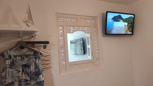 A room with a television displaying a tropical beach scene, a decorative mirror with a geometric frame, and a clothing rack with a tropical print garment. - 50m sandy beach. All comforts ***. Quiet - Private parking - Air-conditioned (Hyères)