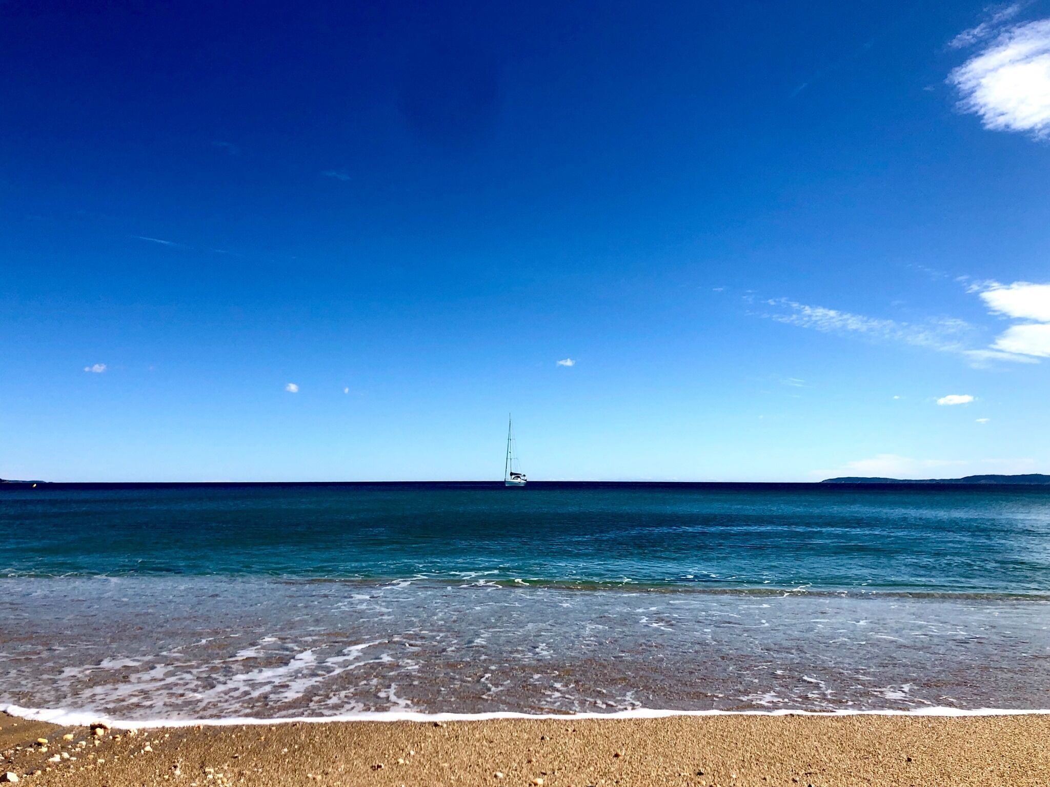 Una playa con cielo despejado, mar tranquilo y un velero a lo lejos.