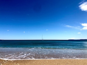 Una platja amb cel clar, mar calma i un veler a la distància.