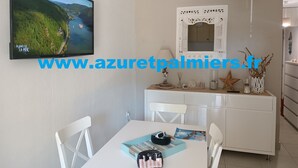A dining area with a white table, chairs, and a white cabinet with decorative items. A television displaying a scenic landscape is mounted on the wall. - 50m sandy beach. All comforts ***. Quiet - Private parking - Air-conditioned (Hyères)