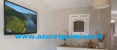 A dining area with a white table, chairs, and a white cabinet with decorative items. A television displaying a scenic landscape is mounted on the wall.