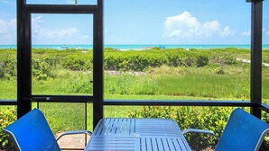 A view from a covered outdoor area with a table and chairs, overlooking a beach and lush greenery.