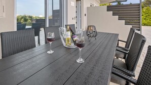 Une table en bois sombre, agrémentée de deux verres à vin et d’une bouteille, entourée de chaises en osier, dans un cadre extérieur offrant une vue sur des bâtiments et de la verdure.