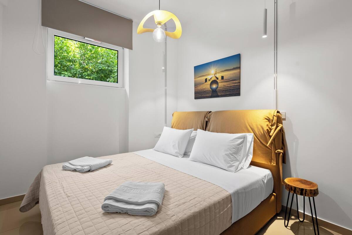 A modern bedroom with a large bed, a window with a view of greenery, a painting of a sunset, and a unique headboard design.