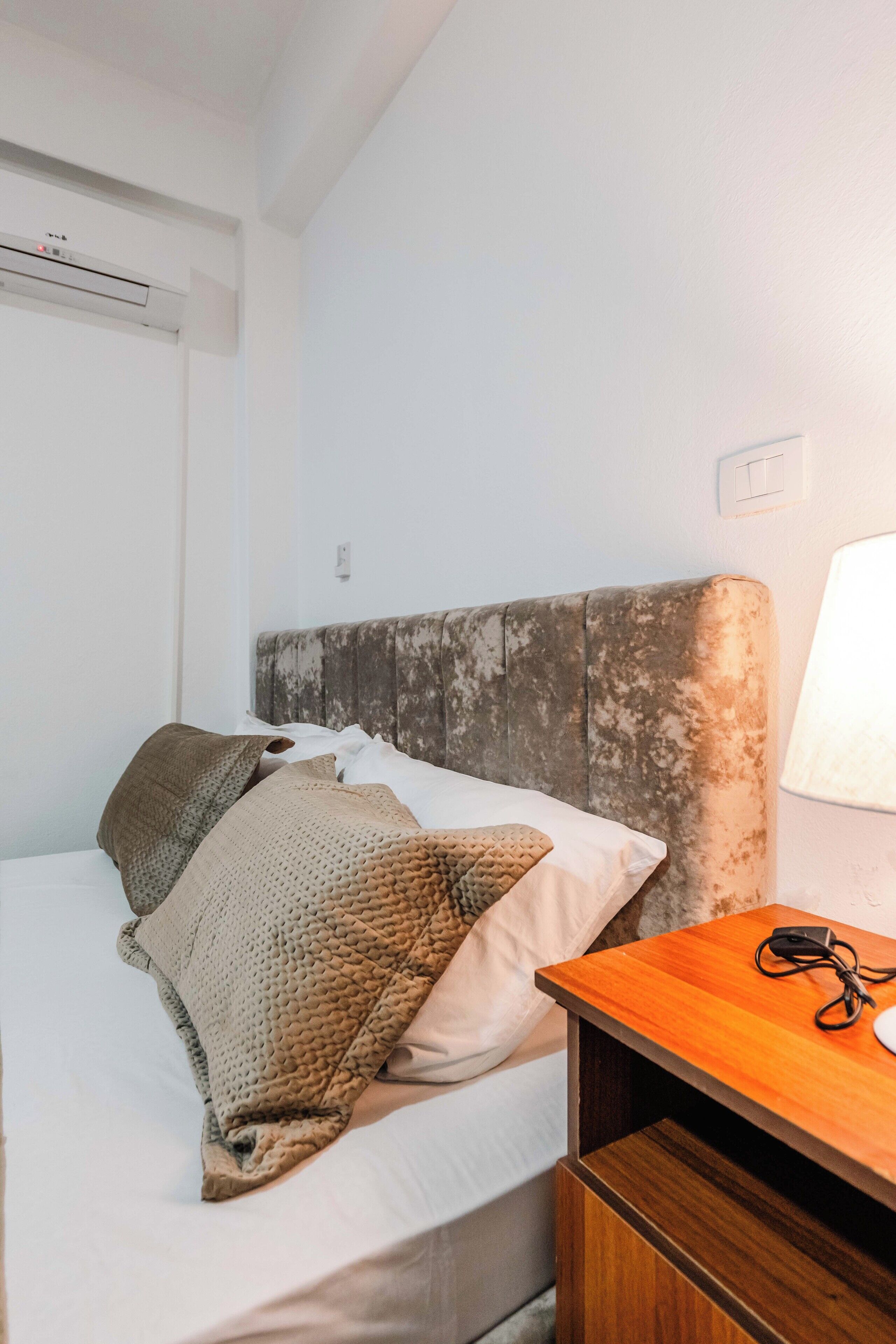 A single bed with a wooden headboard, a bedside table with a lamp, and a power outlet on the wall.