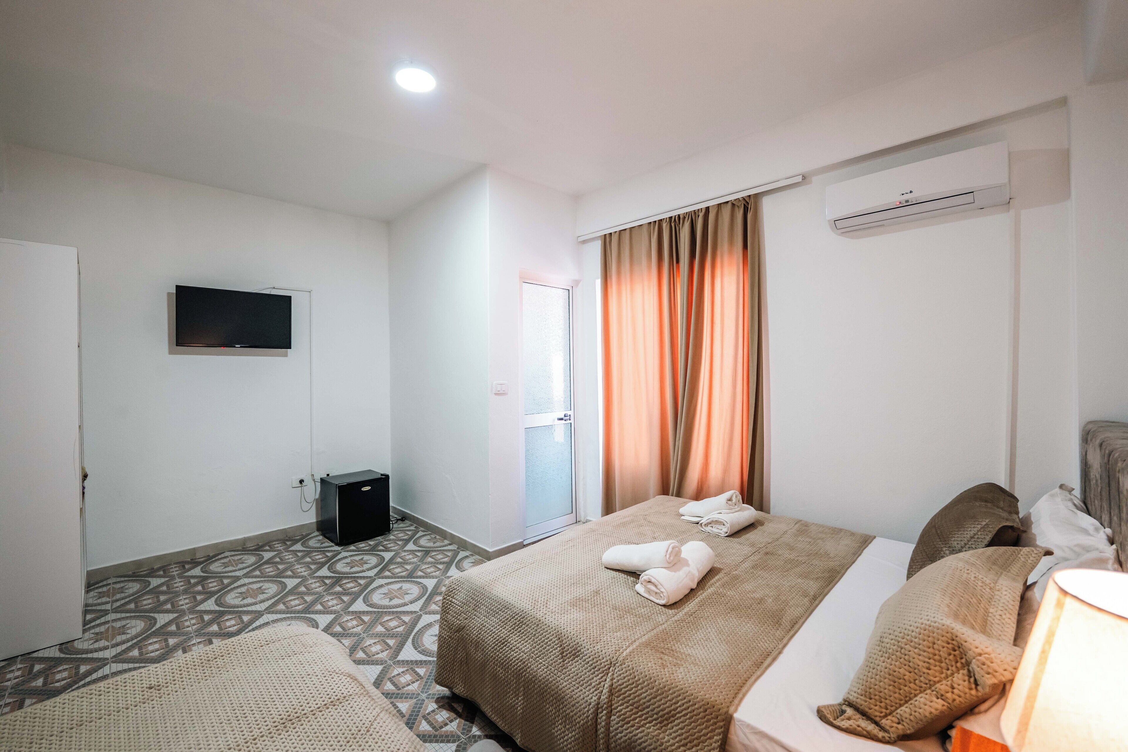 A hotel room with two beds, a television, an air conditioning unit, and patterned floor tiles.