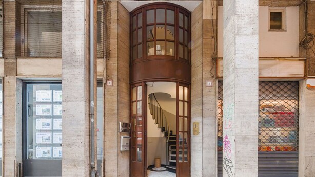 Une entrée d’immeuble avec un escalier en colimaçon, une porte dotée d’un panneau de verre et une fenêtre ornée d’une grille métallique.