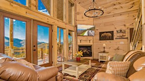 A spacious living room with leather sofas, a coffee table, and a fireplace. Large windows offer a scenic view of the mountains. - 🌲 Paradise View In The Smokies 🌅 Views, Resort Pool & Game Room (Sevierville)