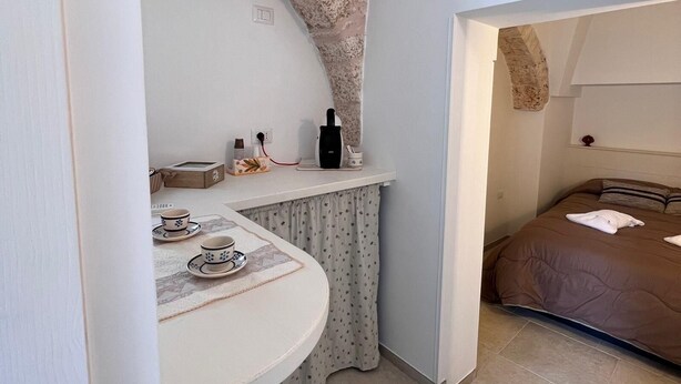 A compact room with a curved white table, a small kitchen area, and a bed with a brown duvet. - Trullo in Light Standard (Alberobello)
