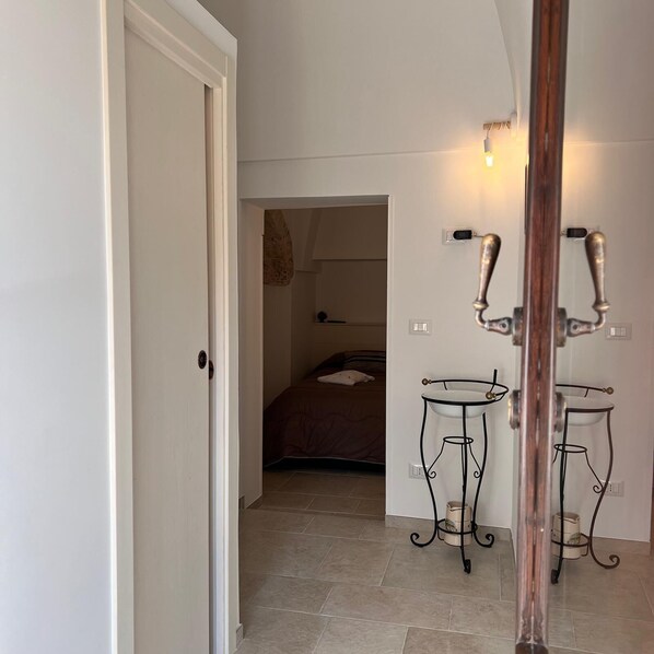 Un couloir avec une porte fermée, un lit dans la pièce adjacente et deux tables en métal, avec un luminaire au-dessus.