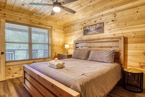 A wooden cabin bedroom with a bed, a nightstand, a lamp, a ceiling fan, and a window with blinds. - Mountain View Cabin Rental-New Cabin-Internet-Hot Tub-Pet Friendly (Sevierville)