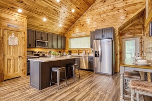 Una cocina tipo cabaña de madera con una isla central, taburetes de bar, electrodomésticos de acero inoxidable y una ventana con persianas.