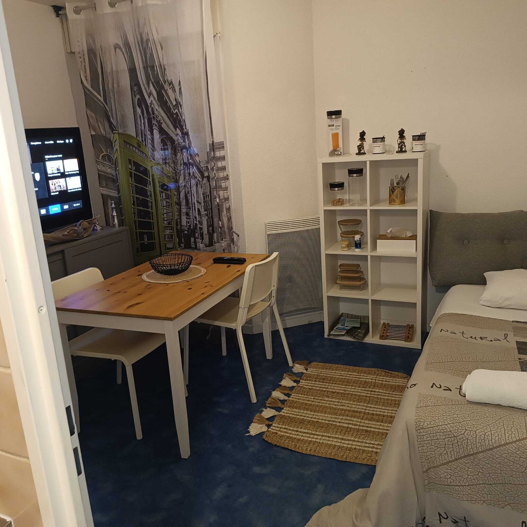 Una habitación con una mesa de madera, sillas blancas, una cama con cubrecama de temática natural, un estante blanco con varios objetos y un mural en la pared con vista a un paisaje urbano.