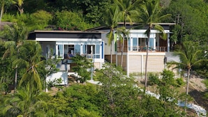 A modern two-story house with large windows, surrounded by lush greenery and palm trees.