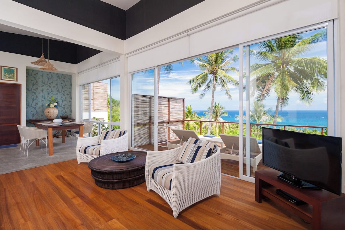 Ein großzügiger Wohnbereich mit einer großen Schiebeglastür, die auf einen Balkon mit Blick auf Palmen und das Meer führt. Es gibt einen Esstisch mit Stühlen, ein Sofa und einen Fernseher auf einem Holzständer.