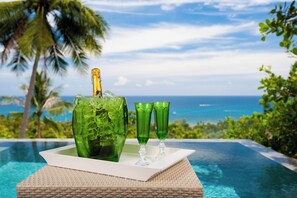 A glass bottle and two glasses on a tray by a pool with a view of the ocean and palm trees. - Overthemoon Luxury Pool Villas (Koh Tao)