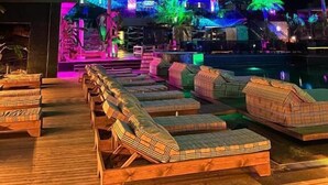 Un edificio de varios pisos con un área de piscina que incluye sillas de descanso y palmeras.