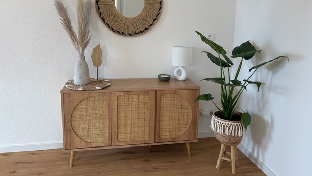 A wooden cabinet with woven panels, a round woven mirror, a white lamp, a potted plant, and decorative items on the cabinet. - GIPFELGLÜCK - Apartment centrally located in Nesselwang - KÖNIGSKARTE  (Nesselwang)