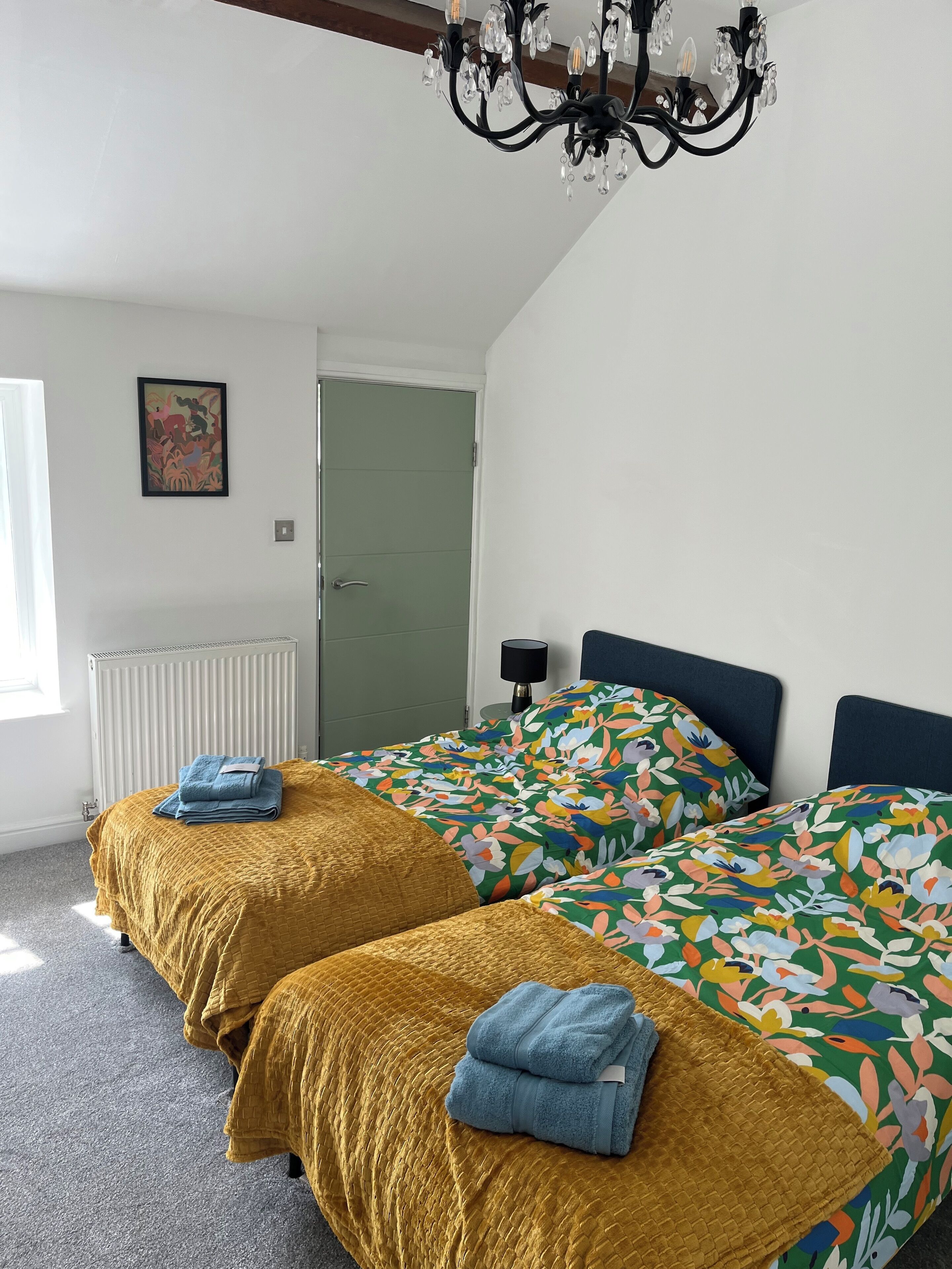 A bedroom with two beds, a radiator, a door, a chandelier, and a wall with a framed picture.