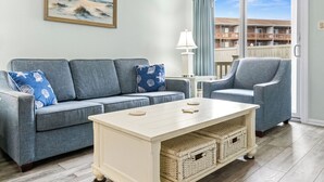 A living room with a grey sofa, a matching armchair, a white coffee table, wicker baskets, a lamp, and a painting of a lighthouse. - North OC gem: 2BR w/ pool, private hot tub & views of Fenwick Lighthouse (Ocean City)