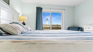 A bedroom with a large bed, a nightstand with a lamp, a chair, and a view of a lighthouse through a window. - North OC gem: 2BR w/ pool, private hot tub & views of Fenwick Lighthouse (Ocean City)
