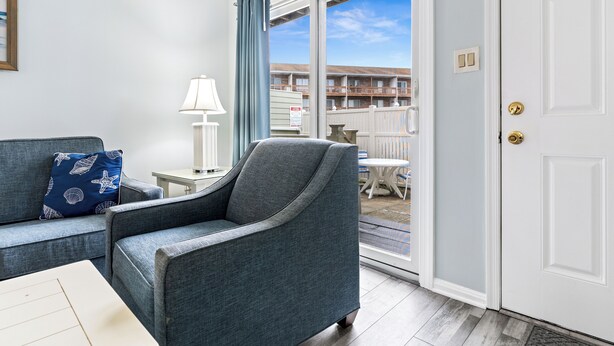 A room with a grey armchair, a blue sofa with starfish pillows, a small white table, and a lamp. There is a sliding glass door leading to an outdoor area with a white fence and a building in the background. - North OC gem: 2BR w/ pool, private hot tub & views of Fenwick Lighthouse (Ocean City)