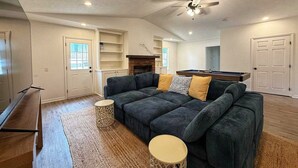 A spacious living room with a dark green sectional sofa, a pool table, and a ceiling fan.