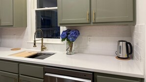 A kitchen with green cabinets, a white countertop, a stainless steel sink, a faucet, a vase with flowers, and a kettle.