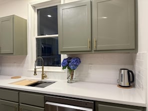 Una cocina con armarios verdes, encimera blanca, pileta de acero inoxidable, grifo, un florero con flores y una tetera.