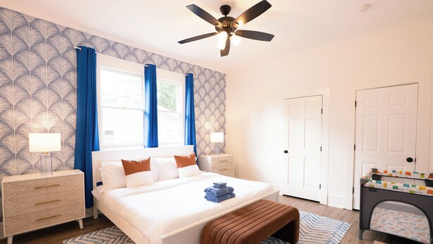 A bedroom with a large bed, a ceiling fan, a dresser, a desk with a chair, and a door.