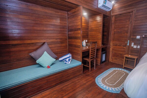 Una habitación pequeña y acogedora con paredes de madera y una cama individual. Hay un banco con almohadones, una bolsa azul, una silla de madera y una mesita con una lámpara.