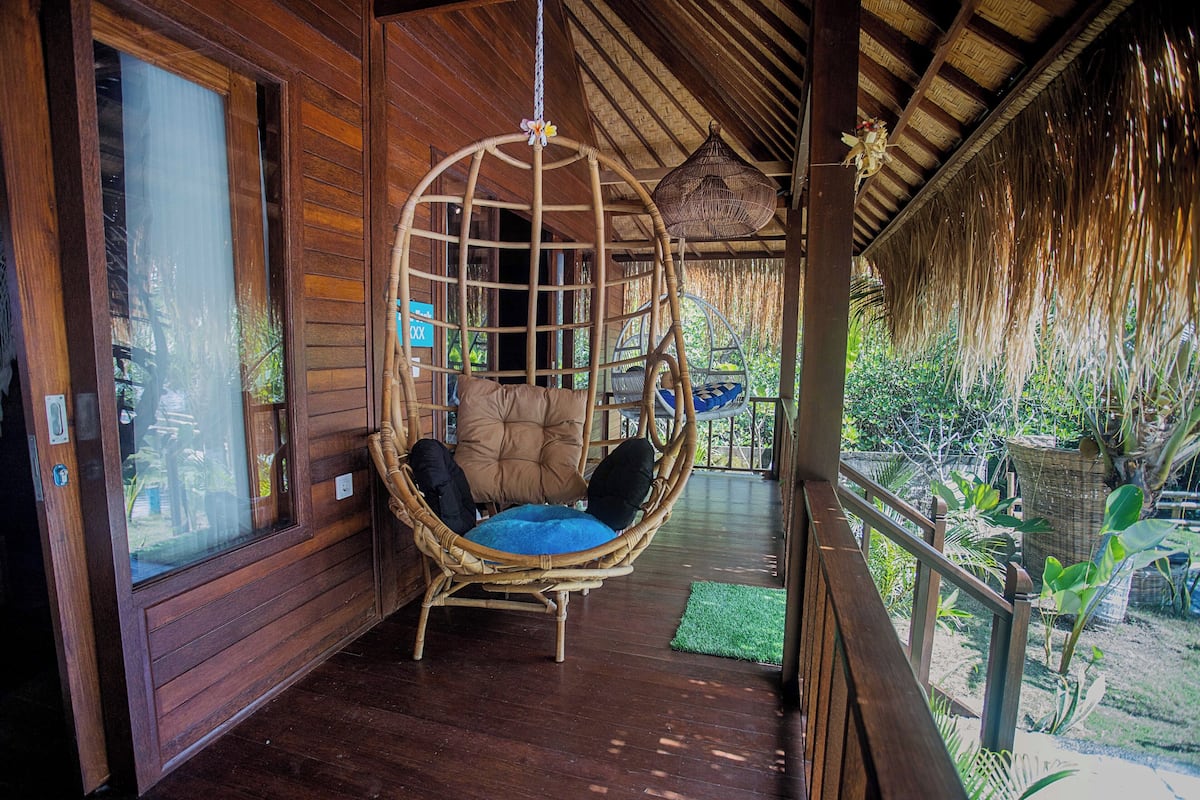 Eine Veranda aus Holz mit einem hängenden Korbstuhl und einem Reetdach.