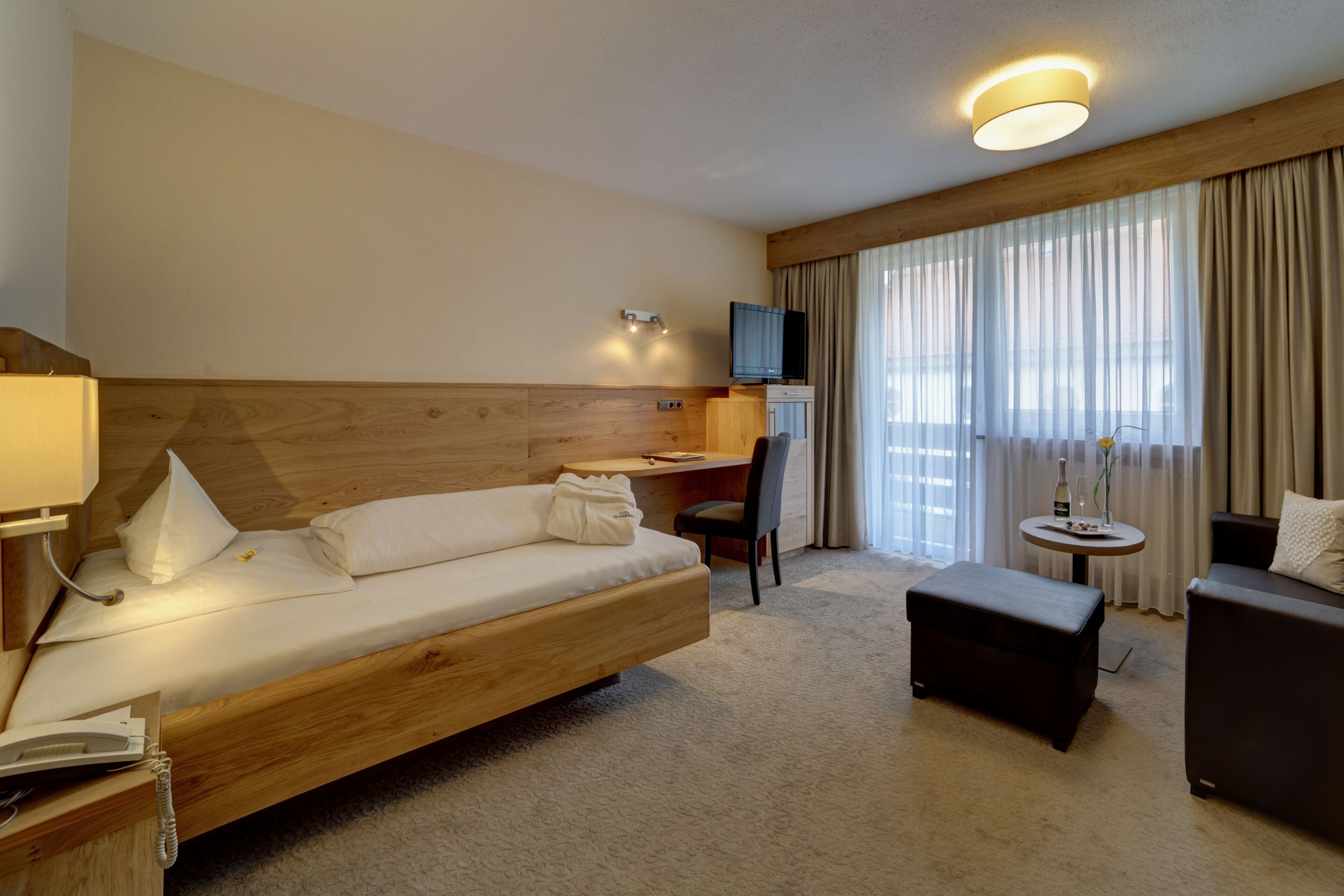 A hotel room with a wooden bed, a desk, a TV, a sofa, and a small round table with a bottle and flowers.