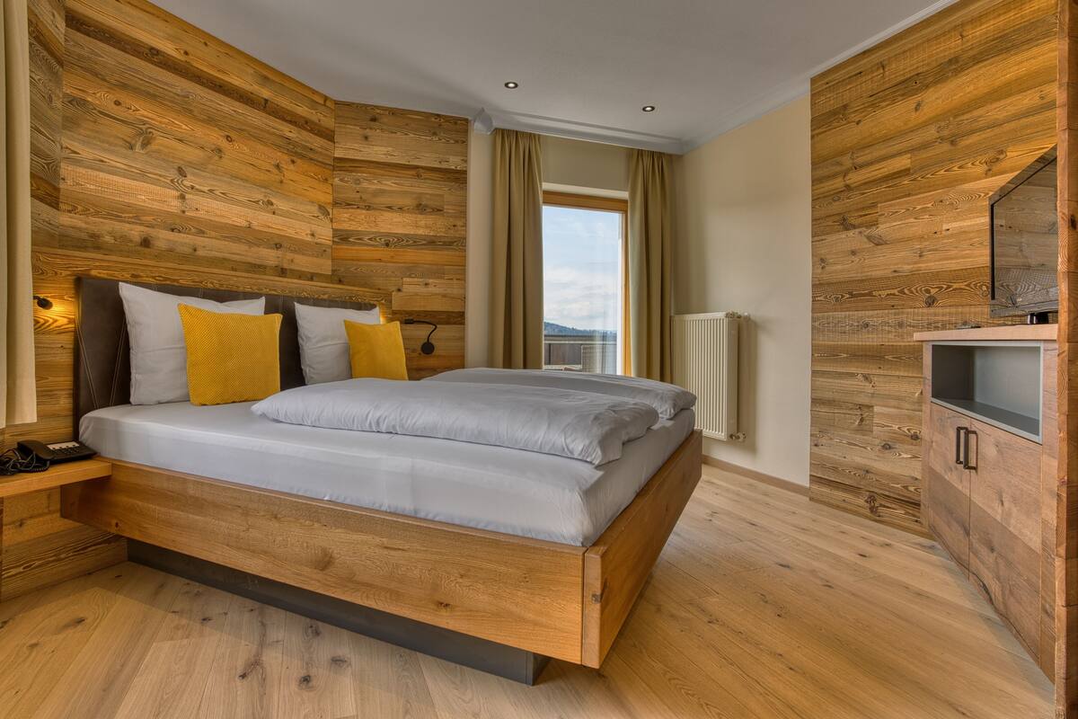 Une chambre à coucher moderne avec une tête de lit en bois, un lit avec une literie blanche et jaune, une table de chevet avec une lampe, et une vue sur l’extérieur par une fenêtre.