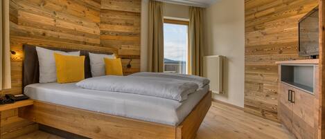 Une chambre à coucher moderne avec une tête de lit en bois, un lit avec une literie blanche et jaune, une table de chevet avec une lampe, et une vue sur l’extérieur par une fenêtre.