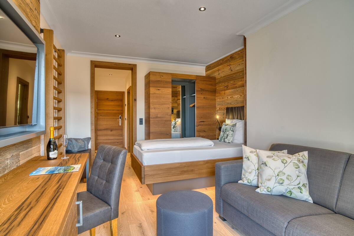 Une chambre moderne avec un plancher en bois, un lit avec un matelas blanc, un canapé gris agrémenté de coussins à motifs, une table en bois avec une bouteille et des verres, et un grand miroir au mur.